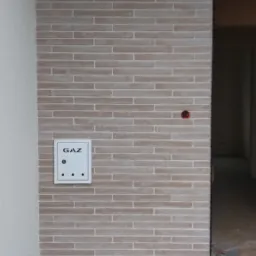 Elewacja budynku wykończona beżową cegłą klinkierową z widoczną skrzynką gazową i wystającą rurką instalacyjną, dolna część elewacji pokryta szarym tynkiem strukturalnym.