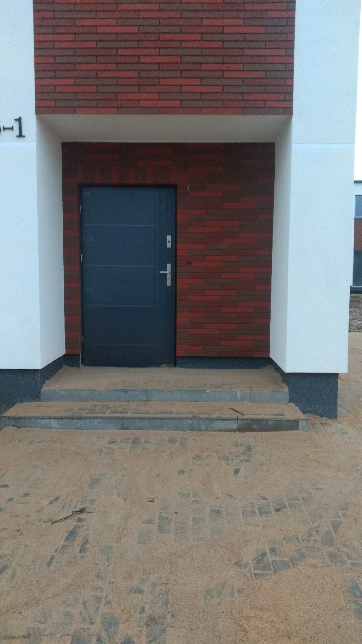 Wejście do budynku z szarą, metalową furtką, wykończone czerwoną cegłą klinkierową i białym tynkiem, z widocznymi schodami wejściowymi i piaskiem na kostce brukowej.