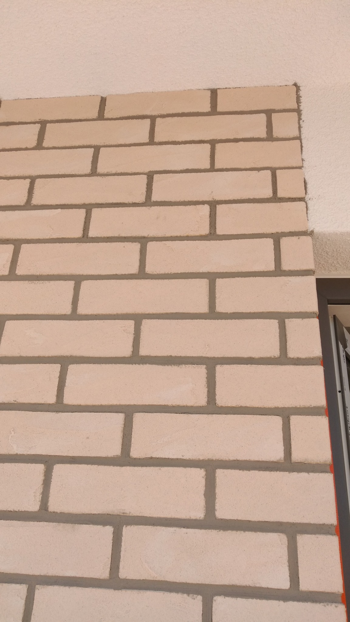 Fragment ściany z jasnej cegły klinkierowej z szarą fugą, widoczny narożnik i fragment okna z szarą ramą, sufit pomalowany na biało z widoczną fakturą.