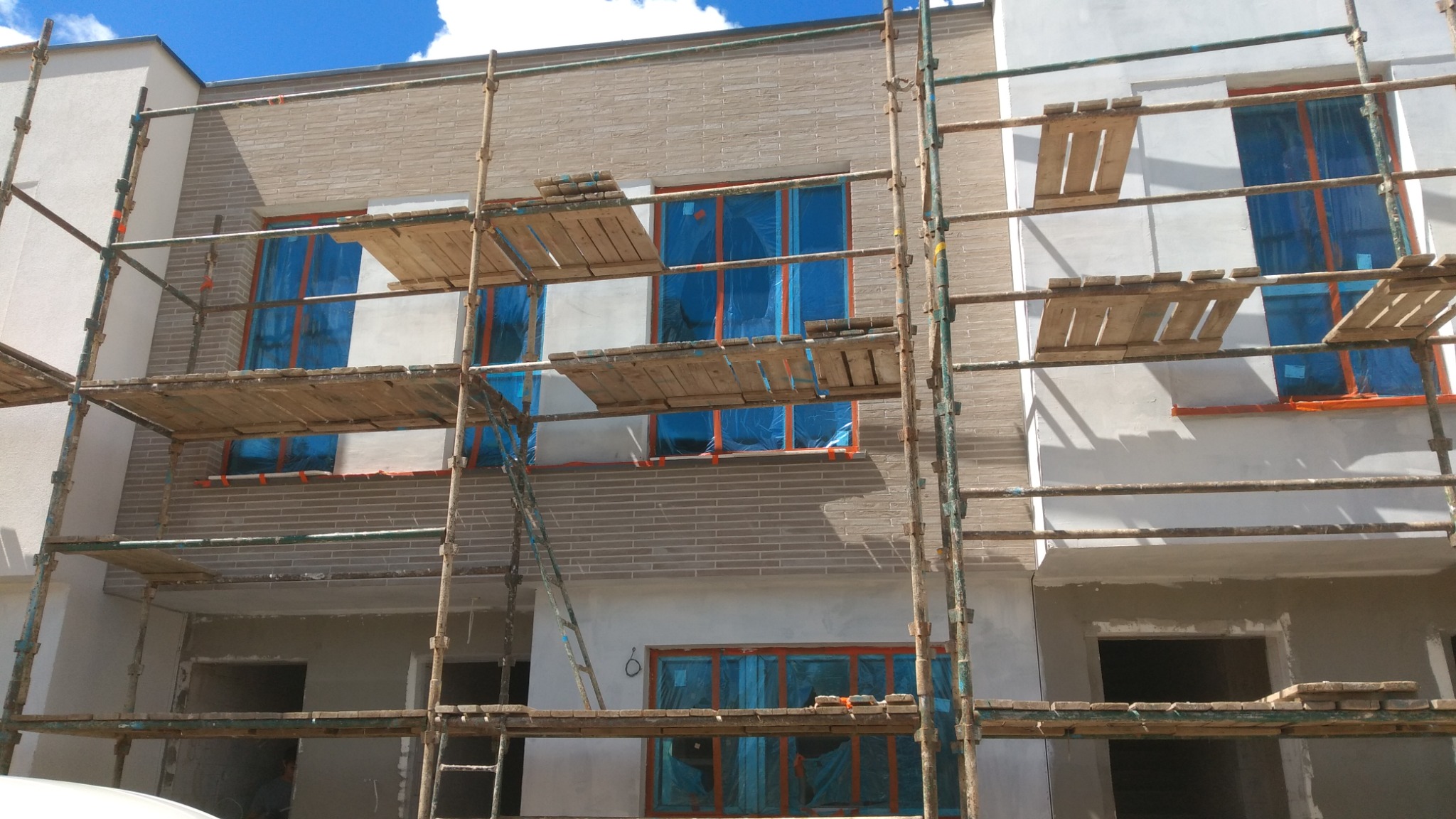 Elewacja budynku w trakcie ocieplania z widocznymi rusztowaniami, oknami zabezpieczonymi folią oraz fragmentami otynkowanej i pomalowanej ściany.