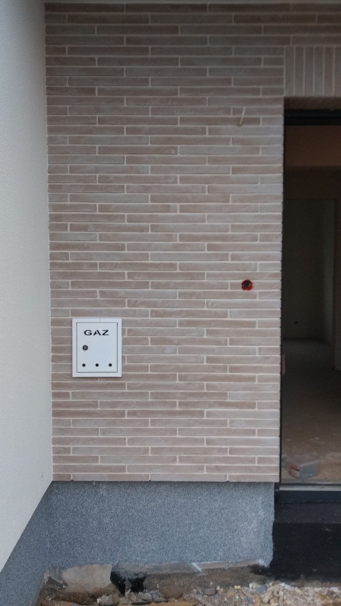 Elewacja budynku wykończona beżową cegłą klinkierową z widoczną skrzynką gazową i wystającą rurką instalacyjną, dolna część elewacji pokryta szarym tynkiem strukturalnym.