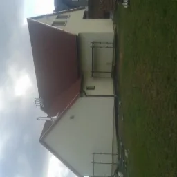 Dom jednorodzinny z nową elewacją i brązowym dachem, widoczny z boku na tle pochmurnego nieba i trawnika, z fragmentami rusztowania.