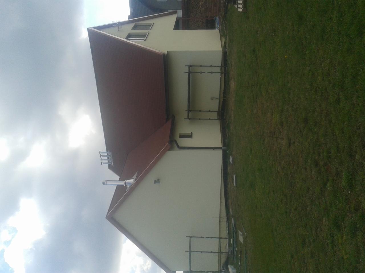 Dom jednorodzinny z nową elewacją i brązowym dachem, widoczny z boku na tle pochmurnego nieba i trawnika, z fragmentami rusztowania.