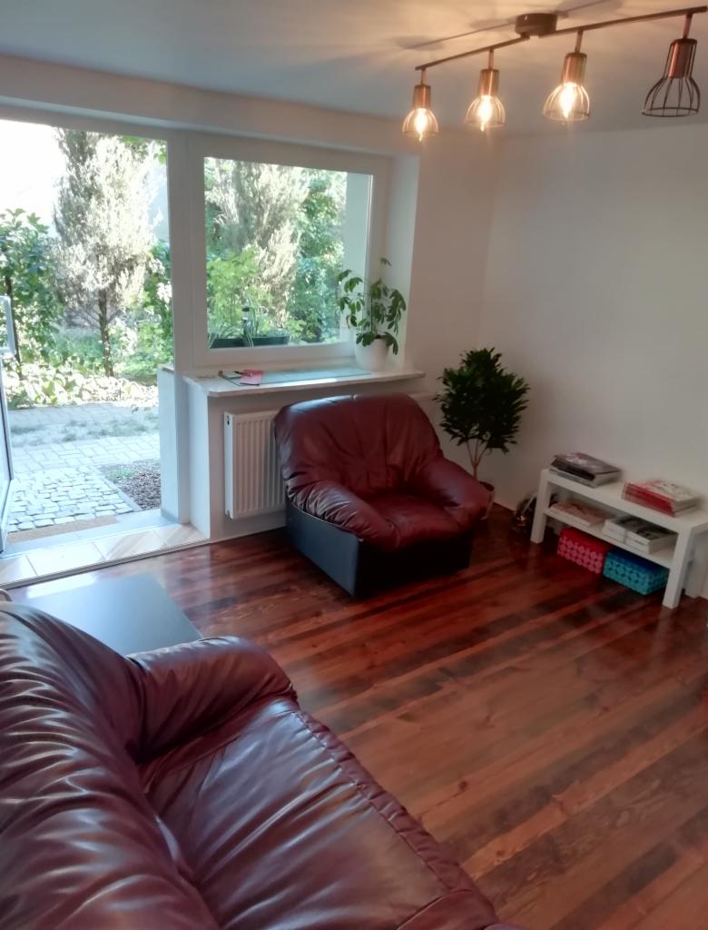 Częściowy widok salonu z brązową skórzaną sofą, fotelem, drewnianą podłogą i oknem z widokiem na zieleń.