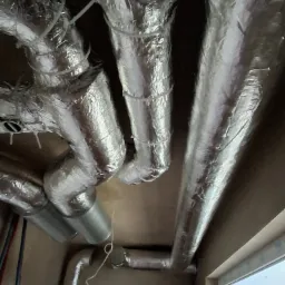 Srebrne, izolowane kanały wentylacyjne pod sufitem, częściowo w folii aluminiowej, z widocznymi elementami mocującymi i fragmentem okna w tle.
