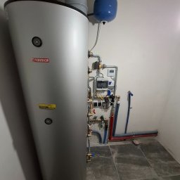 EVENLIGHT Marcin Sobierajski - Pionowy, srebrny zbiornik Termica z podłączonymi rurami miedzianymi i niebieskim zbiornikiem wyrównawczym, zainstalowany w pomieszczeniu z szarą podłogą imitującą marmur.