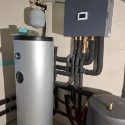 Szary zasobnik ciepłej wody użytkowej Kensol z podłączonymi rurami w izolacji, obok stoi szary zasobnik Proton. Widoczne elementy instalacji i manometr.