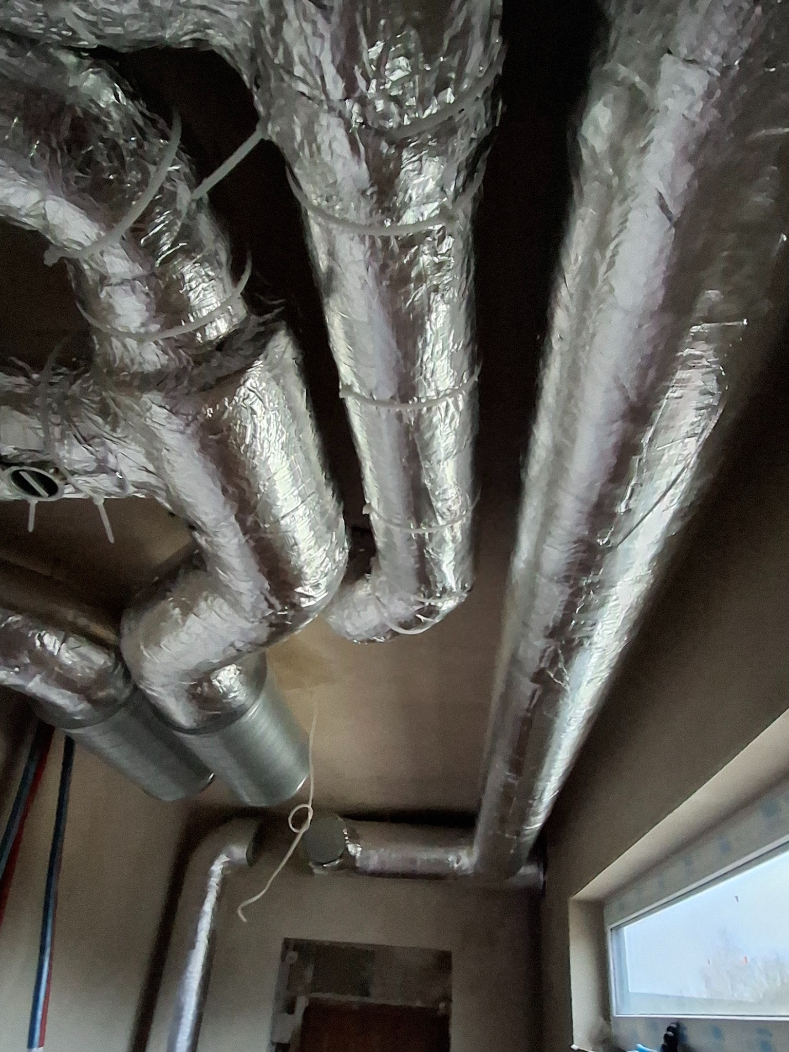 Srebrne, izolowane kanały wentylacyjne pod sufitem, częściowo w folii aluminiowej, z widocznymi elementami mocującymi i fragmentem okna w tle.