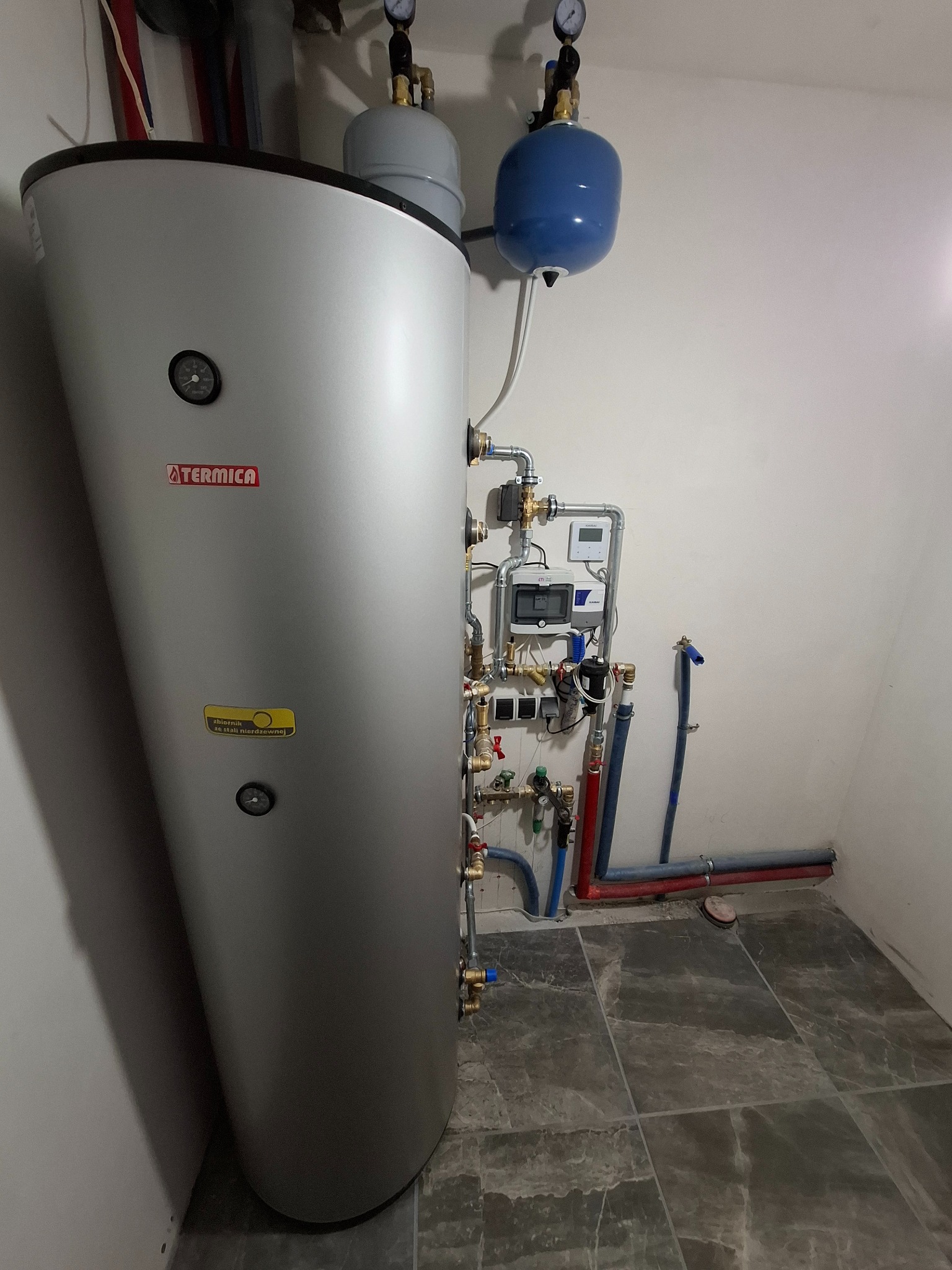 Pionowy, srebrny zbiornik Termica z podłączonymi rurami miedzianymi i niebieskim zbiornikiem wyrównawczym, zainstalowany w pomieszczeniu z szarą podłogą imitującą marmur.