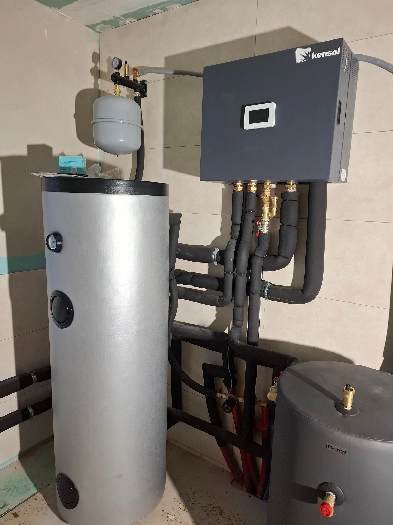 Szary zasobnik ciepłej wody użytkowej Kensol z podłączonymi rurami w izolacji, obok stoi szary zasobnik Proton. Widoczne elementy instalacji i manometr.