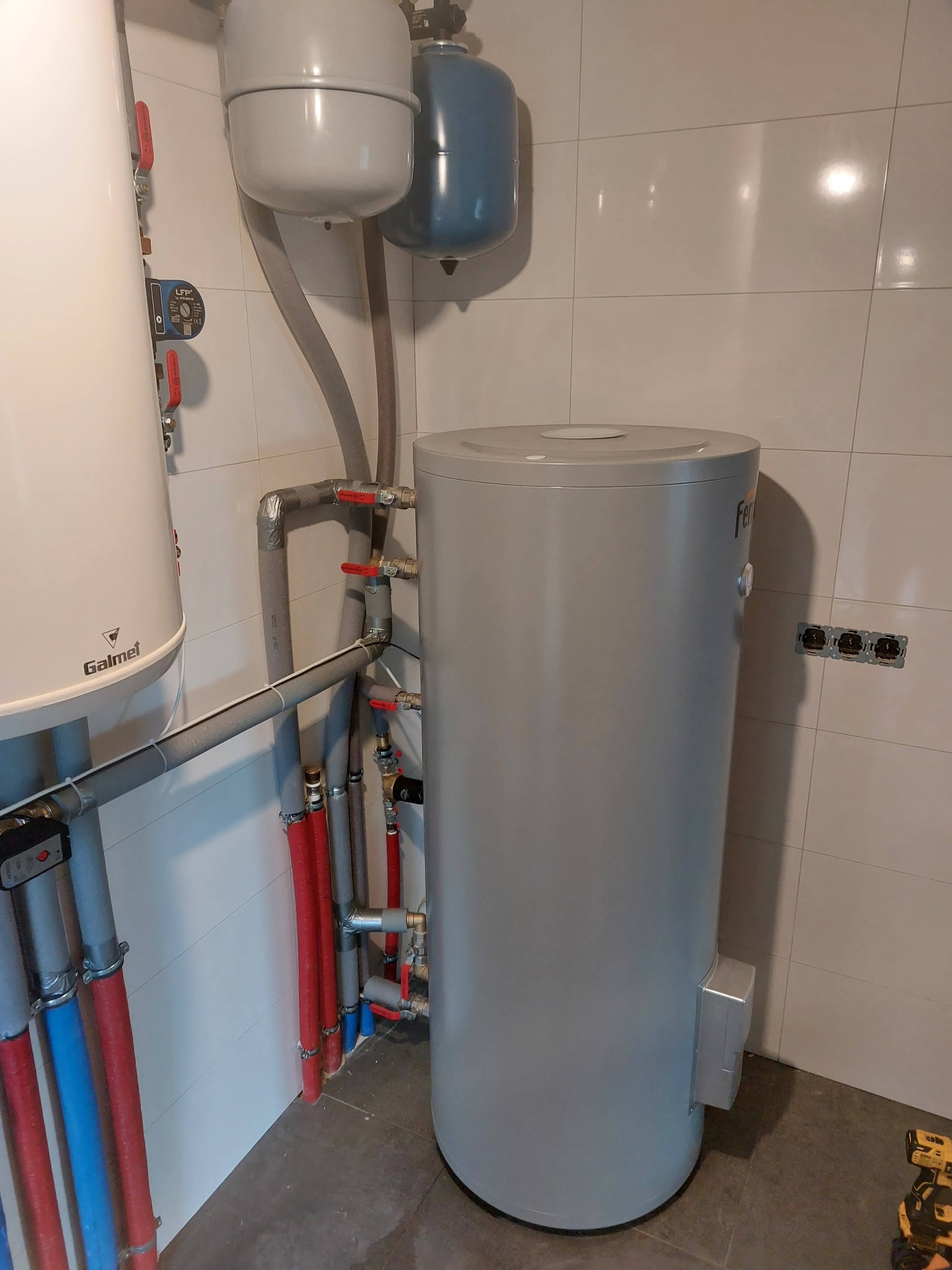Szary, cylindryczny zasobnik ciepłej wody Ferroli z podłączeniami rur miedzianych i izolowanych, widoczne zawory kulowe z czerwonymi rączkami, obok biały zbiornik Galmet, tło stanowią białe płytki...