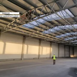 Wnętrze nowo wybudowanej, przestronnej hali z konstrukcji stalowej z oknami dachowymi, widoczny pracownik w odblaskowej kamizelce i mokra posadzka.