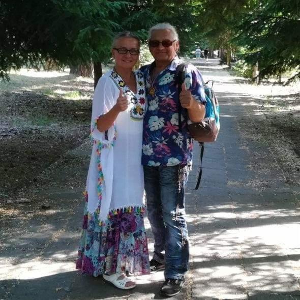 Para w parku, oboje z podniesionymi kciukami, kobieta w białej sukience z kolorowymi zdobieniami, mężczyzna w kwiecistej koszuli i okularach przeciwsłonecznych, z plecakiem na ramieniu.