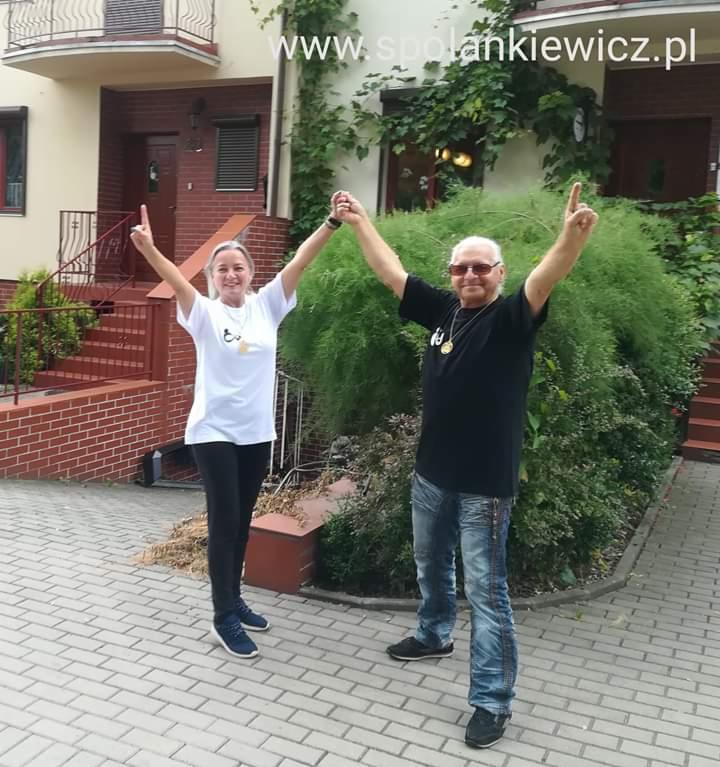 Uśmiechnięta kobieta i mężczyzna w okularach, trzymający się za ręce i wskazujący palcami do góry, na tle budynku z cegły i zieleni