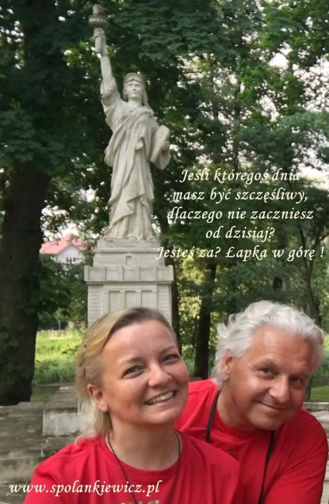 Uśmiechnięta para na tle pomnika Statuy Wolności z sentencją o szczęściu i adresem strony internetowej w dolnej części kadru.