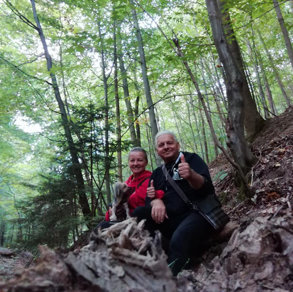 Para z psem siedzi na powalonym pniu drzewa w lesie, oboje pokazują kciuki w górę.