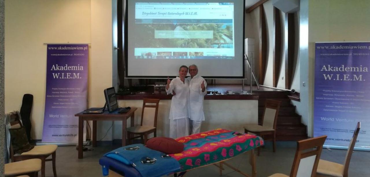 Para w białych strojach pozuje przed ekranem projekcyjnym z wyświetloną stroną internetową gabinetu terapii naturalnych, obok leżanka do masażu z kolorowym pokrowcem i banery reklamowe.