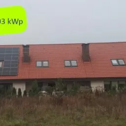 Dom jednorodzinny z czerwoną dachówką, na której zamontowane są panele słoneczne o mocy 6.03 kWp, widoczne okno dachowe pod panelami, pochmurne niebo w tle.