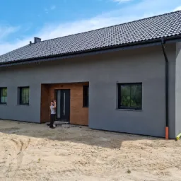 Nowoczesny dom jednorodzinny z szarą elewacją, czarną dachówką i rynnami, drewnianymi elementami przy wejściu, sfotografowany w słoneczny dzień z widocznym fragmentem ogrodu i niezagospodarowanym...