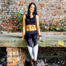 Uśmiechnięta kobieta w sportowym stroju, eksponująca wyrzeźbiony brzuch, siedzi na betonowej konstrukcji na tle ceglanej ściany z zielenią.