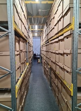 Magazyn z rzędami wysokich regałów wypełnionych kartonowymi pudłami, widok z wąskiego korytarza między regałami, w głębi widoczny wózek transportowy.