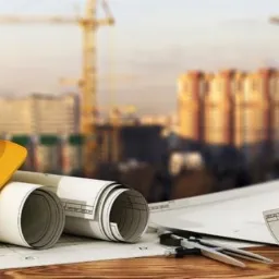 Żółty kask budowlany, zwinięte plany architektoniczne, cyrkiel i ołówek na tle rozmytego krajobrazu miejskiego z dźwigami budowlanymi.