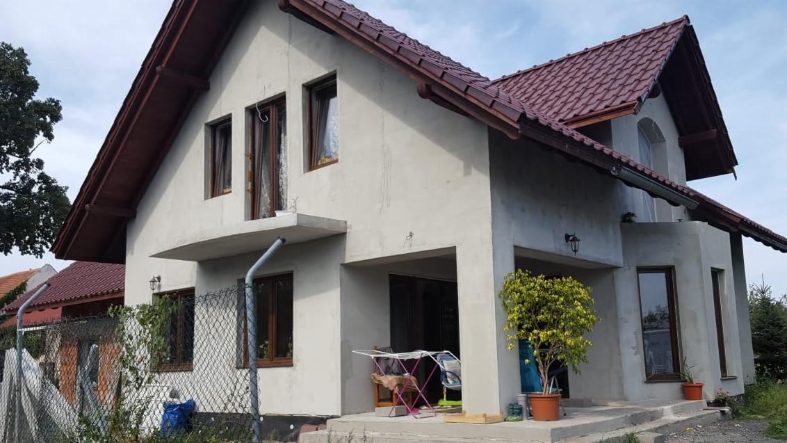 Dom jednorodzinny z szarą elewacją w trakcie wykańczania, widoczne okna z brązowymi ramami, czerwony dach, rynna i drzewko w doniczce przy wejściu.