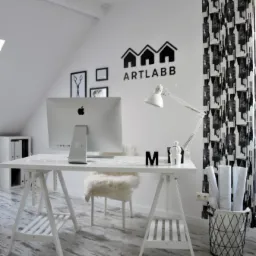 Białe biuro w skandynawskim stylu z biurkiem na kozłach, komputerem iMac, krzesłem z futerka i czarno-białymi zasłonami. Na ścianie logo firmy ART LABB.