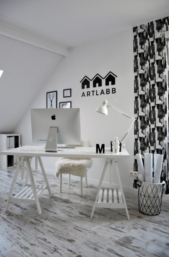 Białe biuro w skandynawskim stylu z biurkiem na kozłach, komputerem iMac, krzesłem z futerka i czarno-białymi zasłonami. Na ścianie logo firmy ART LABB.