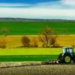 Zielony traktor z podpiętym pługiem na polu uprawnym, w tle widoczne zielone pola i błękitne niebo z chmurami.
