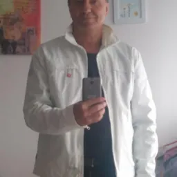 Mężczyzna w jasnej kurtce robi selfie w lustrze; w tle widoczne obrazy na ścianie i fragment pokoju.