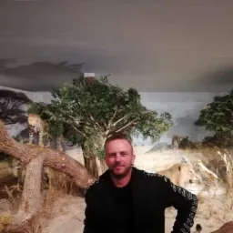 Mężczyzna pozuje na tle dioramy z afrykańskimi zwierzętami: lwy, gepardy i drzewa. Ekspozycja muzealna z oświetleniem.