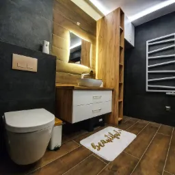 Nowoczesna łazienka z ciemnymi płytkami imitującymi kamień, drewnianymi akcentami, podświetlanym lustrem, wiszącą toaletą, grzejnikiem drabinkowym i dywanikiem z napisem 'beautiful'.