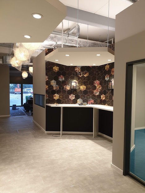 Wnętrze komercyjne po remoncie: ściana z heksagonalną tapetą, biały blat, oświetlenie punktowe i wiszące lampy kuliste, widok na plac budowy przez przeszklone drzwi.