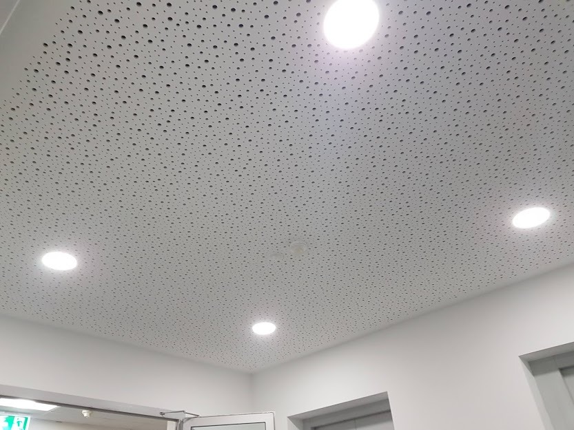 Biały sufit podwieszany z perforacją i wbudowanymi okrągłymi lampami LED, widok z dołu pod kątem w jasnym wnętrzu.