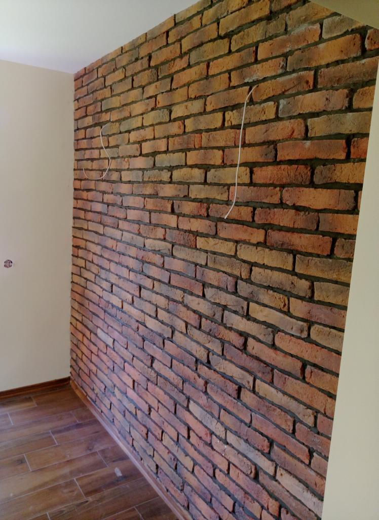 Ściana wyłożona cegłą klinkierową w odcieniach czerwieni i brązu, z widocznymi przewodami elektrycznymi wystającymi ze ściany, drewnopodobne panele na podłodze.