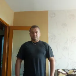 Mężczyzna w Bolesławcu stoi na tle ściany z wzorem róż, obok pomalowanej na beżowo ściany i drzwi. Widoczny fragment sufitu i sofy.