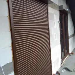 Brązowa roleta zewnętrzna zamontowana na ścianie z białych bloczków, widoczne niedokończone prace wykończeniowe i narzędzia.