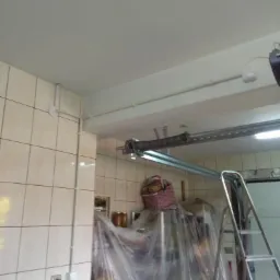 Białe rury elektryczne natynkowe prowadzone po ścianie z płytek w garażu, widoczna instalacja bramy garażowej i drabina aluminiowa.