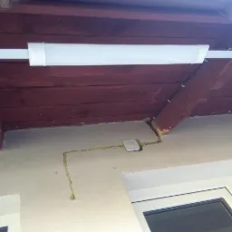 Zamontowana biała lampa LED w korytarzu pod drewnianym sufitem, widoczne przewody elektryczne w listwach i puszka instalacyjna na ścianie.