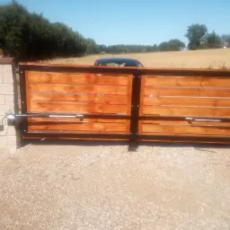 Automatyczna brama przesuwna z drewnianymi panelami, widoczne siłowniki, sterownik i lampa ostrzegawcza na słupku, w tle pole i drzewa.