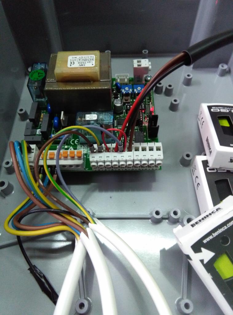 Wnętrze szarej skrzynki sterowniczej z podłączonymi przewodami o różnych kolorach do listwy zaciskowej, widoczny transformator i elementy elektroniki, częściowo widoczne dwa białe moduły sterujące...