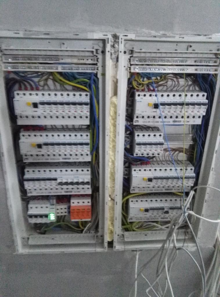 Dwa rozdzielnice elektryczne w trakcie montażu, widoczne liczne przewody, bezpieczniki i wyłączniki, szara ściana w tle, widoczne ślady montażu na ścianie.