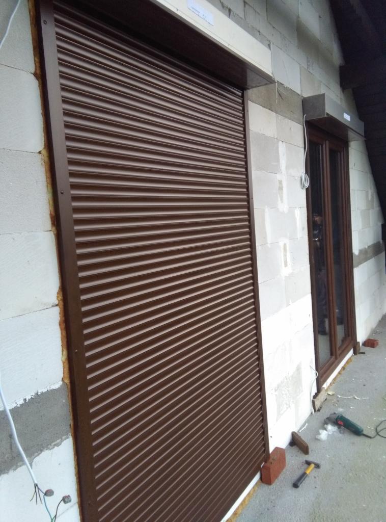 Brązowa roleta zewnętrzna zamontowana na ścianie z białych bloczków, widoczne niedokończone prace wykończeniowe i narzędzia.