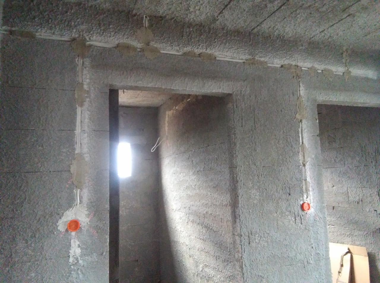 Surowy stan budynku w trakcie prac elektrycznych: widoczne przewody przytwierdzone do sufitu i ścian, puszka instalacyjna w kolorze pomarańczowym na ścianie, otwór okienny w tle.