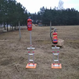 Dwa czerwone gaśnice na metalowych stojakach z pomarańczowymi podstawami, ustawione na trawiastym terenie z drzewami w tle.