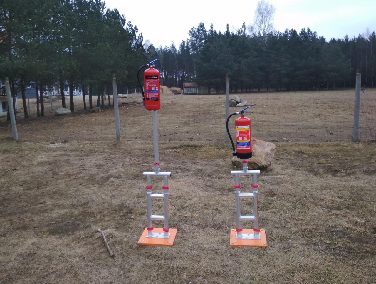 Dwa czerwone gaśnice na metalowych stojakach z pomarańczowymi podstawami, ustawione na trawiastym terenie z drzewami w tle.