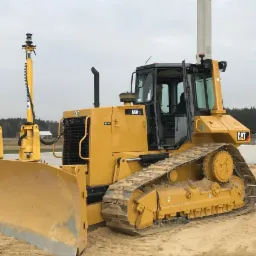 Żółty spychacz Caterpillar D6M z systemem niwelacji laserowej na placu budowy, widoczne betonowe elementy konstrukcyjne w tle.