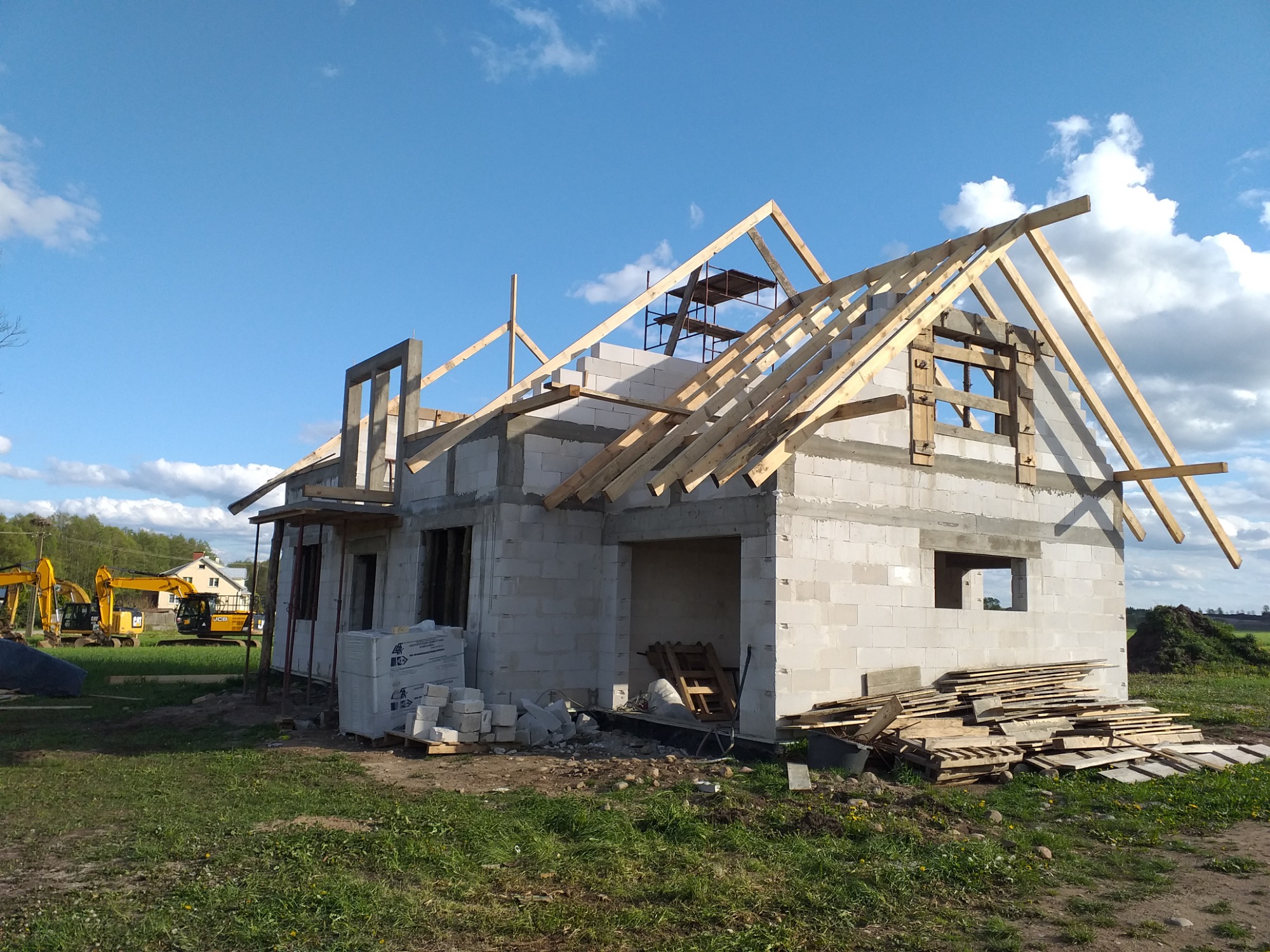 Budowa domu jednorodzinnego w stanie surowym otwartym: ściany z betonu komórkowego, drewniana konstrukcja dachu, widoczne rusztowania i maszyny budowlane w tle.