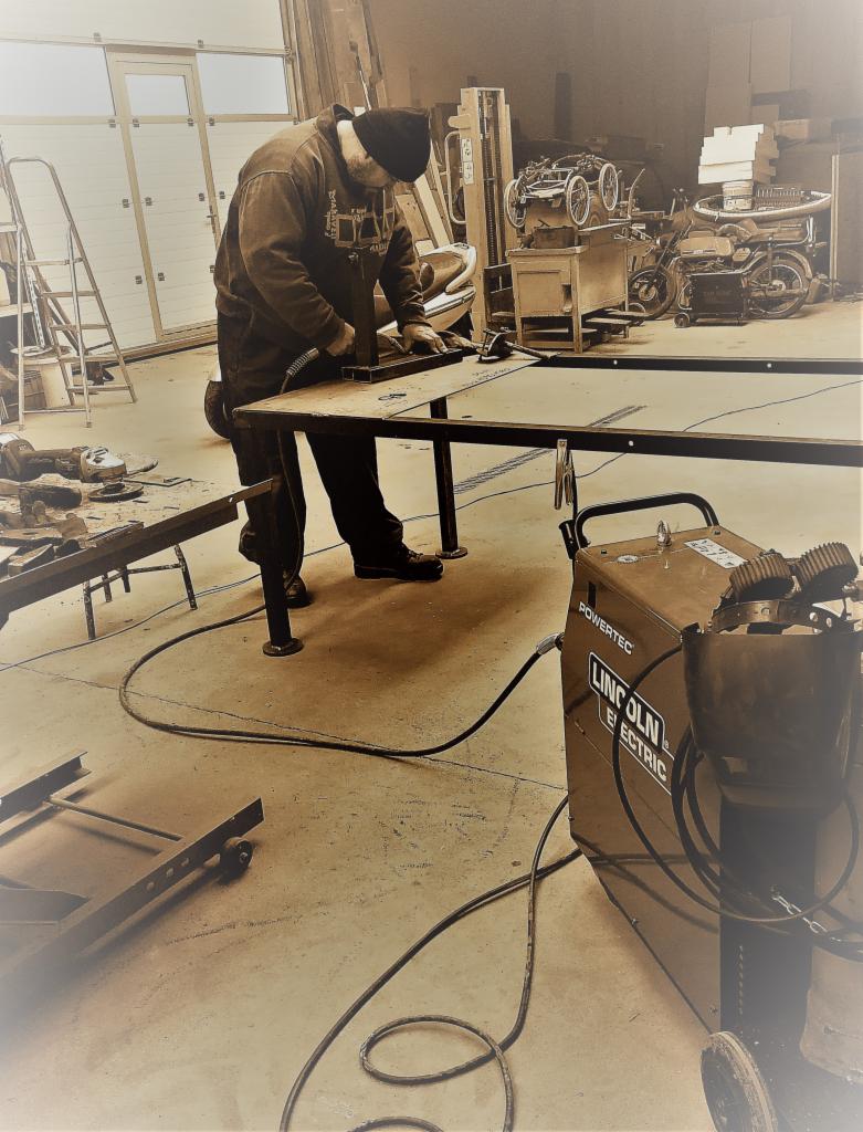 Pracownik w czapce, w warsztacie, obrabia metalowy profil na stole roboczym, widoczny spawarka Lincoln Electric i narzędzia.